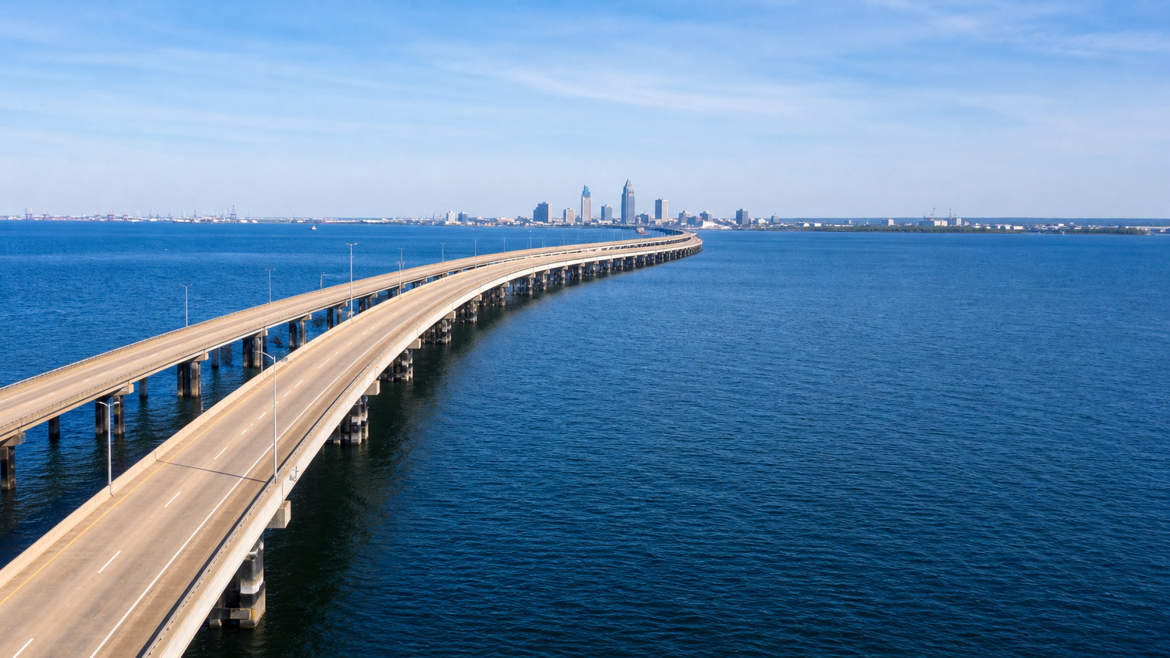 I-10 Bayway bridge crossing Mobile Bay Alabama on a sunny day, wide concrete bridge spanning blue bay water with Mobile city skyline faint in the distance