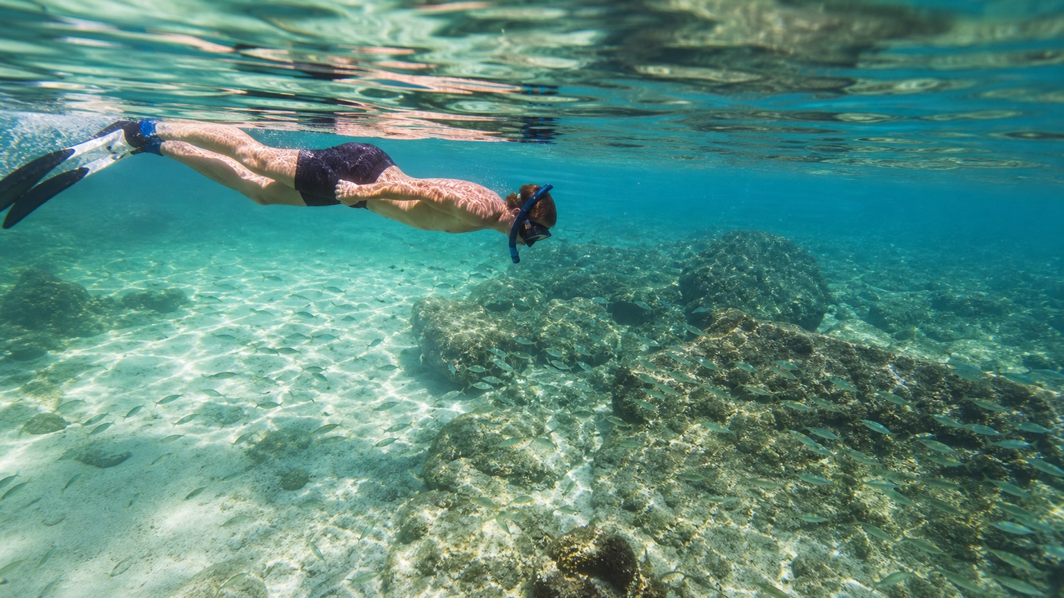 Snorkeler floating face-down in crystal-clear turquoise water at the Destin jetties, looking down at fish swimming around submerged rock structure